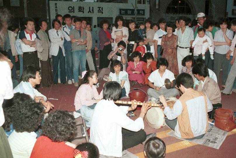 8. 1985년 5월 12일, 대학로 마당놀이 축제에서 둥글게 앉아 악기를 연주하는 사람들. 주변 참가자들도 함께 둘러서서 듣고 있다.